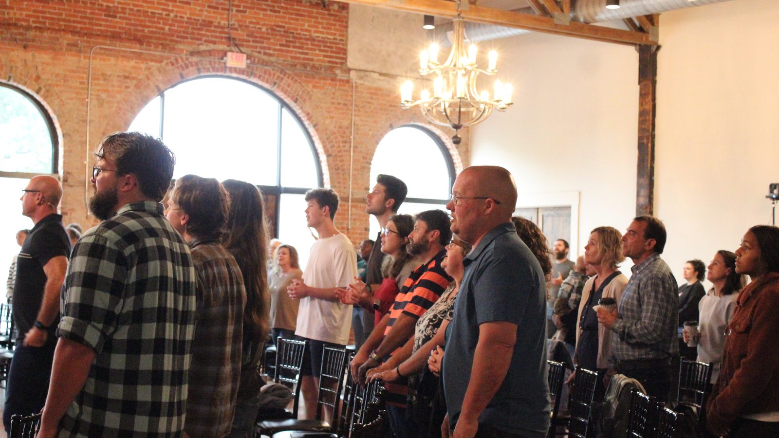 A packed congregation of men, women, and teenagers standing shoulder to shoulder in worship inside a brick-and-timber sanctuary