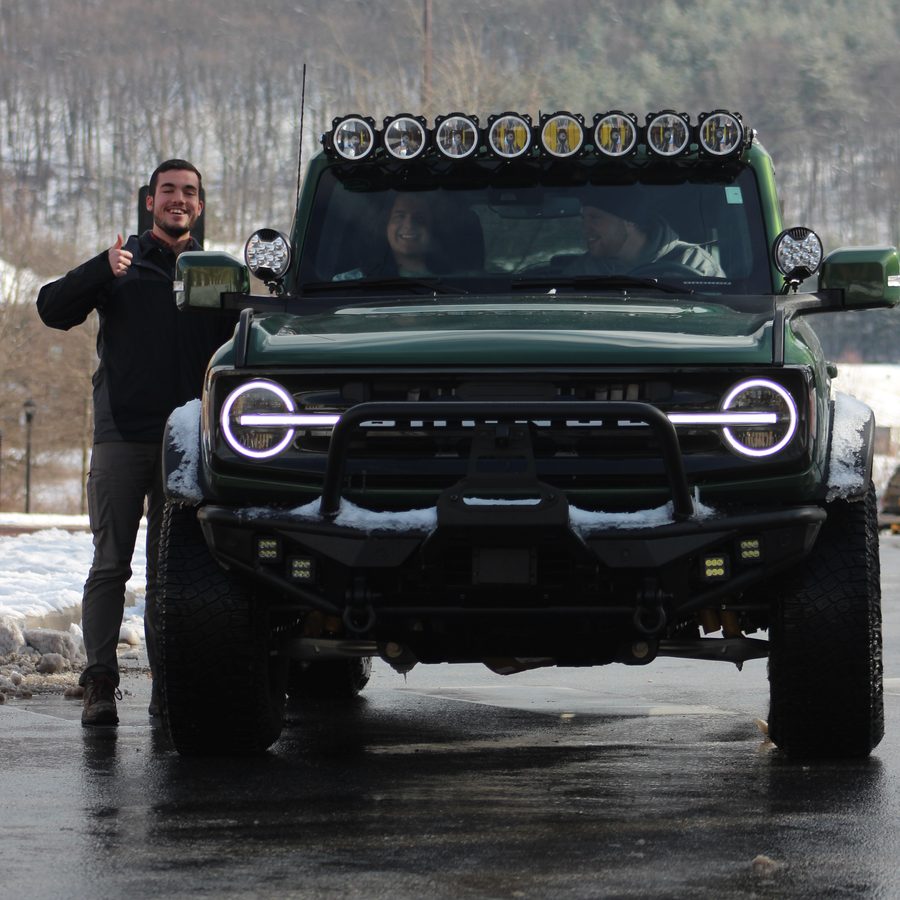 A young man gives a thumbs up beside a lifted green Ford Bronco with two friends smiling inside, on a snowy winter afternoon